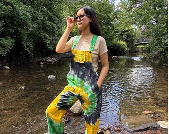 May include: Tie-dye overalls in yellow, green, and black. The overalls have a bib pocket and green straps. The model is wearing a light-colored t-shirt and black sandals. The logo "A. Fashion" is in the top left corner.