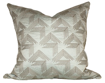 May include: A square decorative pillow with a light blue background and a geometric pattern in a darker, muted tone. The pattern consists of stitched lines forming arrow-like shapes across the pillow's surface. The pillow has a soft, textured appearance.