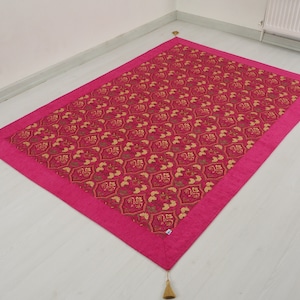 Könnte beinhalten: Eine rosa Decke mit goldfarbenem Muster und Quasten an jeder Ecke.