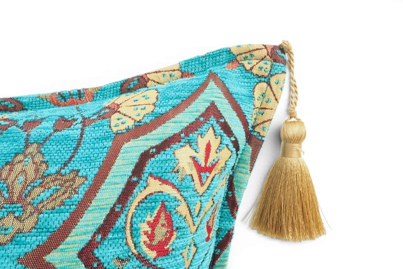 May include: A decorative throw pillow corner featuring a vibrant turquoise fabric with a woven pattern in shades of cream, brown, and red. The corner is finished with a single, long, light gold-colored tassel attached by a twisted cord.