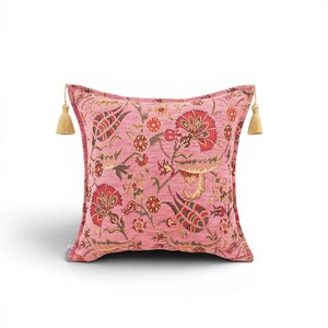 May include: A square throw pillow in a vibrant pink hue featuring a detailed floral pattern in shades of red, green, and gold. The pillow is finished with a textured fabric and includes a decorative beige tassel attached to each top corner.