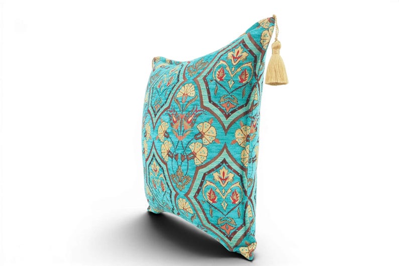May include: A square throw pillow featuring a vibrant turquoise background with a repeating pattern of cream-colored floral motifs outlined in dark brown. The pillow is accented with a single beige tassel attached to the top corner of the fabric.