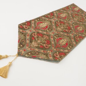 May include: A green table runner with a floral pattern in red, gold, and pink. The runner has tassels on each end.