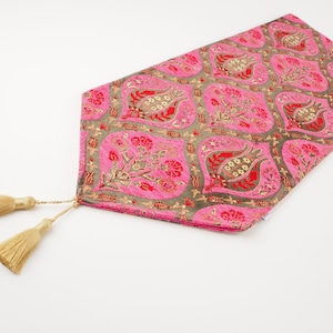 Puede incluir: Un camino de mesa rosa y dorado con un patrón floral y borlas. El camino de mesa está hecho de tejido tejido y tiene un diseño tradicional.