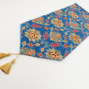 Op de afbeelding: Een blauwe tafelrunner met een bloemenpatroon en gouden kwastjes. De runner heeft een traditioneel ontwerp met rode, gele en groene bloemen.
