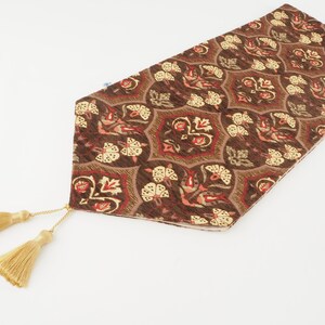 May include: A decorative table runner with a brown and gold floral pattern. The runner has a repeating design of stylized flowers and leaves. Gold tassels hang from the end of the runner. The runner is made of a textured fabric.