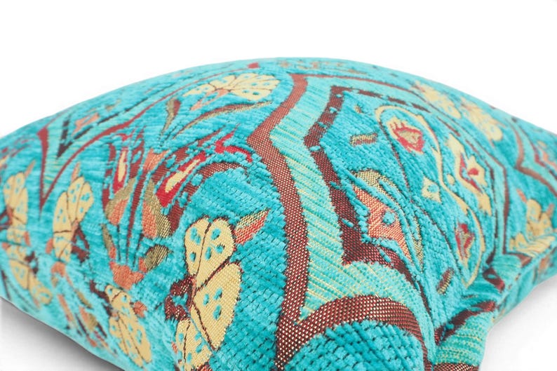 May include: A square throw pillow featuring a vibrant turquoise velvet base with a woven pattern of stylized flowers and geometric shapes in shades of cream, orange, and deep red. The fabric has a textured, raised pile finish throughout.