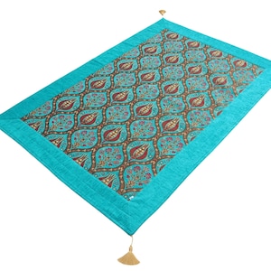 May include: A rectangular turquoise table runner with a detailed floral pattern in teal, burgundy, and brown. The runner has a turquoise border and gold tassels at each corner. Home decor.