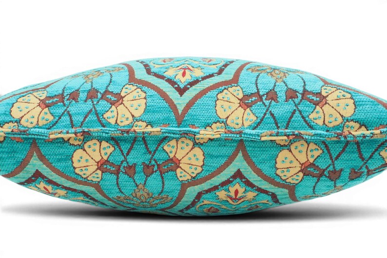May include: A rectangular throw pillow featuring a vibrant turquoise fabric base with a repeating pattern of cream-colored floral motifs and dark brown geometric lines. The pillow has a visible piped edge seam running horizontally across the center.