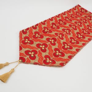 May include: Red and gold table runner with a floral pattern and tassels on the ends.