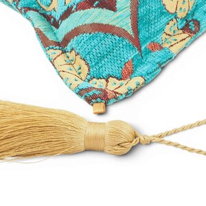 May include: A corner of a turquoise throw pillow featuring a woven floral pattern in cream and dark red, attached to a light gold decorative tassel with a braided loop. The fabric has a textured, raised appearance with visible thread details.
