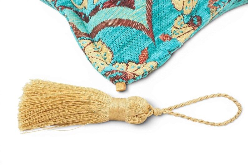 May include: A corner of a turquoise throw pillow featuring a woven floral pattern in cream and dark red, attached to a light gold decorative tassel with a braided loop. The fabric has a textured, raised appearance with visible thread details.