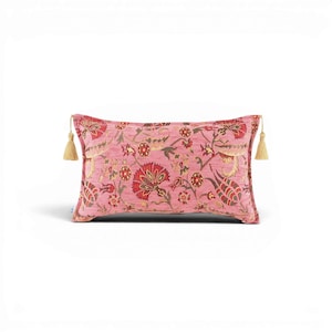 May include: A rectangular throw pillow in a dusty pink hue featuring a woven floral pattern in shades of red, green, and gold. The pillow is finished with small, light-colored decorative tassels attached to each of the two side corners.