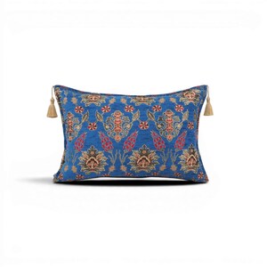 Op de afbeelding: Een rechthoekig blauw sierkussen met een bloemenpatroon in rood, bruin en goud. Het kussen heeft aan elk uiteinde een kwastje. Het patroon bevat gestileerde bloemen en geometrische vormen, waardoor een traditioneel ontwerp ontstaat.