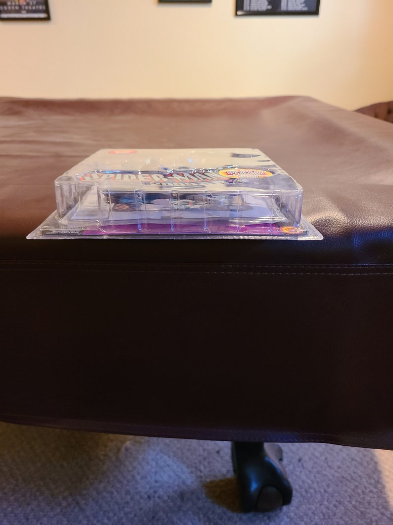 May include: A clear plastic package containing a Spider-Man action figure. The package has a purple and white background with the words "Spider-Man" printed in red and blue. The action figure is visible through the plastic and is wearing a red and blue suit.