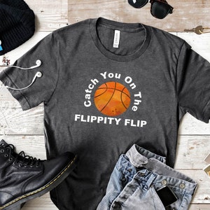 May include: A dark gray t-shirt with a white graphic of a basketball with the text "Catch You On The Flippity Flip".