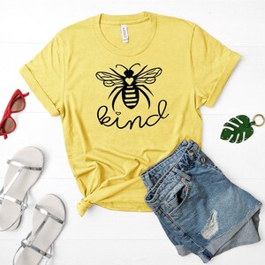 May include: Yellow t-shirt featuring a black bee graphic and the word "kind" printed on it. The bee has a small heart on its body.