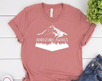 May include: A dusty rose t-shirt with a white mountain and trees graphic and the text "Adventure Awaits". The t-shirt is laid out on a white wooden surface with a pair of white trainers and blue jeans.