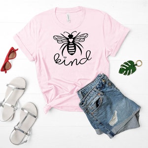 May include: Pink t-shirt featuring a black and white bee graphic and the word "kind" printed on it. The shirt is styled with denim shorts and silver sandals.