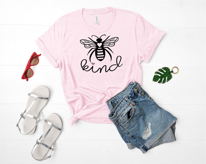 May include: Pink t-shirt featuring a black and white bee graphic and the word "kind" printed on it. The shirt is styled with denim shorts and silver sandals.