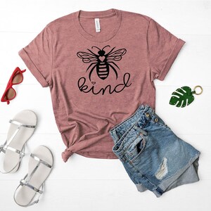 May include: A mauve t-shirt with a black bee graphic and the word "kind" printed on the front. The bee has a heart in its body. The shirt is laid out on a white surface with a pair of silver sandals, a pair of red sunglasses, and a pair of blue denim shorts.