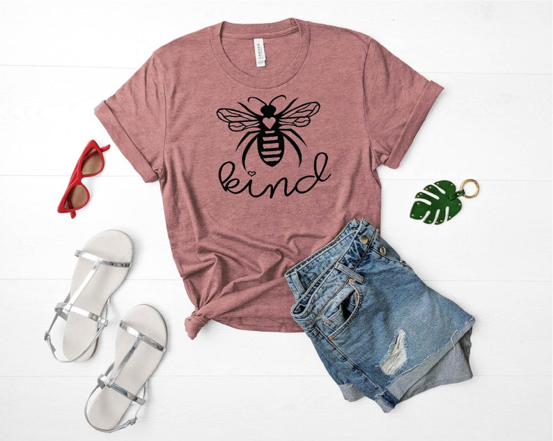 May include: A mauve t-shirt with a black bee graphic and the word "kind" printed on the front. The bee has a heart in its body. The shirt is laid out on a white surface with a pair of silver sandals, a pair of red sunglasses, and a pair of blue denim shorts.