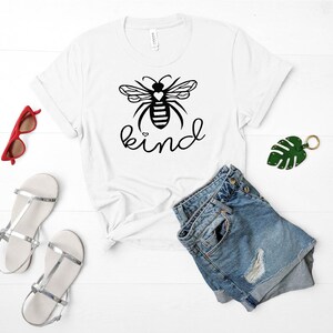 May include: White t-shirt featuring a black and white bee graphic with the word "kind" printed in black. The bee has a small heart on its body.