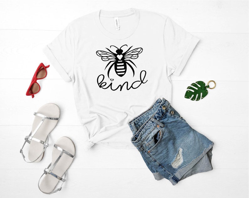 May include: White t-shirt featuring a black and white bee graphic with the word "kind" printed in black. The bee has a small heart on its body.