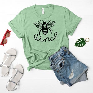 May include: A green t-shirt featuring a black bee graphic and the word "kind" printed on it. The bee has a small heart on its body. The shirt is laid out on a white surface with a pair of denim shorts, a pair of white sandals, and a pair of red sunglasses.