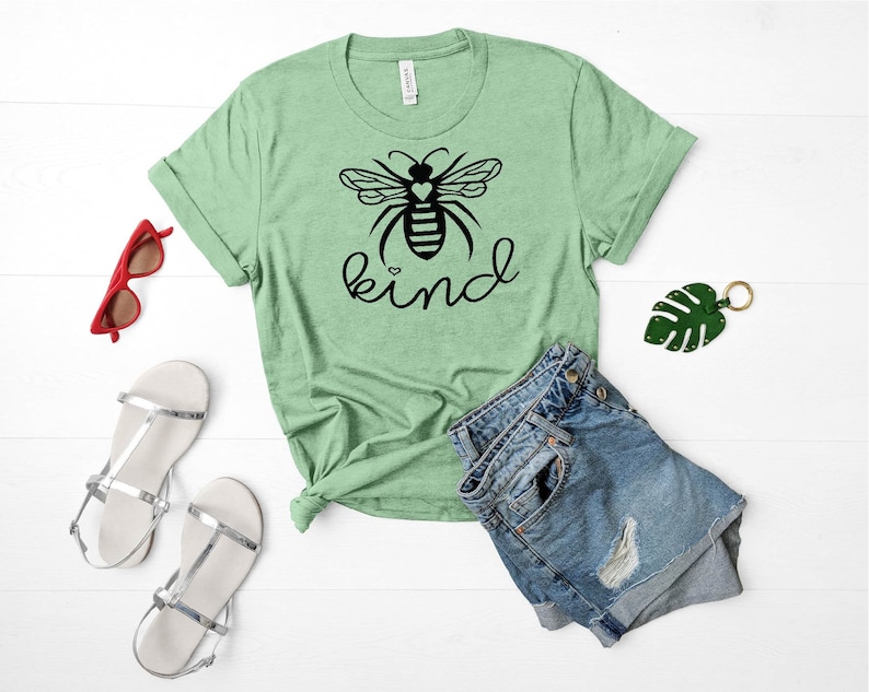 May include: A green t-shirt featuring a black bee graphic and the word "kind" printed on it. The bee has a small heart on its body. The shirt is laid out on a white surface with a pair of denim shorts, a pair of white sandals, and a pair of red sunglasses.
