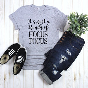 Puede incluir: Una camiseta gris con el texto "It's Just a Bunch of Hocus Pocus" impreso en negro. La camiseta se combina con jeans azules y zapatillas negras.