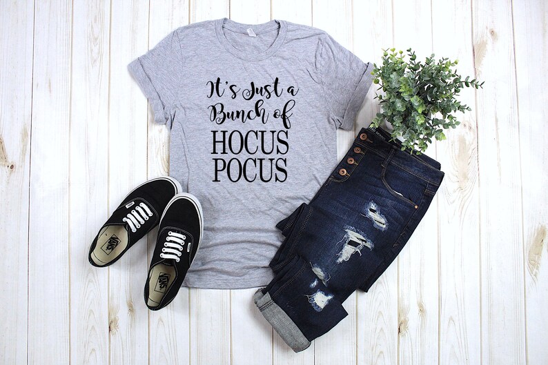 Puede incluir: Una camiseta gris con el texto "It's Just a Bunch of Hocus Pocus" impreso en negro. La camiseta se combina con jeans azules y zapatillas negras.
