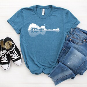 May include: A teal blue t-shirt with a white graphic of a guitar made of trees and a reflection in water. The t-shirt is laid out on a white wooden surface with a pair of black canvas shoes and blue jeans.