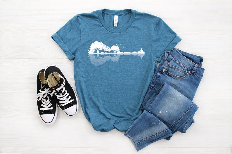 May include: A teal blue t-shirt with a white graphic of a guitar made of trees and a reflection in water. The t-shirt is laid out on a white wooden surface with a pair of black canvas shoes and blue jeans.