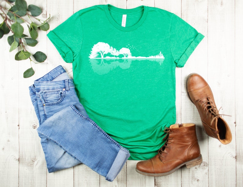 May include: A green t-shirt with a white graphic of a guitar silhouette with trees reflected in the water. The graphic is a reflection of a guitar with trees in the background.