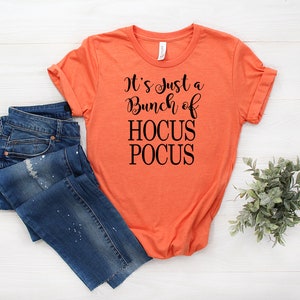 Puede incluir: Camiseta naranja con texto negro que dice "It's Just a Bunch of Hocus Pocus".