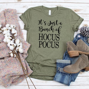 Puede incluir: Una camiseta verde con el texto "It's just a bunch of HOCUS POCUS" en letras negras. La camiseta se muestra sobre un fondo de madera blanca con un par de botines marrones de tobillo, unos vaqueros azules y un su&eacute;ter de punto rosa y blanco.