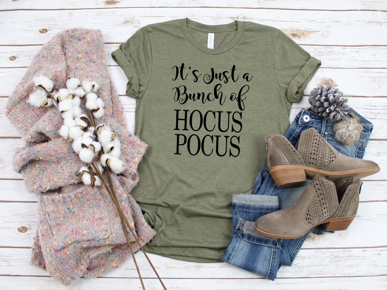Puede incluir: Una camiseta verde con el texto "It's just a bunch of HOCUS POCUS" en letras negras. La camiseta se muestra sobre un fondo de madera blanca con un par de botines marrones de tobillo, unos vaqueros azules y un su&eacute;ter de punto rosa y blanco.