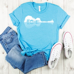 May include: A light blue t-shirt with a white graphic of a guitar silhouette with trees reflected in water. The t-shirt is laid out on a white wooden surface with a pair of white canvas shoes and blue jeans.
