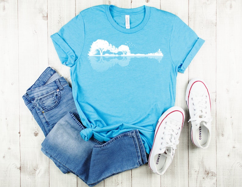 May include: A light blue t-shirt with a white graphic of a guitar silhouette with trees reflected in water. The t-shirt is laid out on a white wooden surface with a pair of white canvas shoes and blue jeans.
