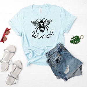 May include: A light blue t-shirt featuring a black bee graphic and the word "kind" printed in black. The bee has a small white heart on its body.