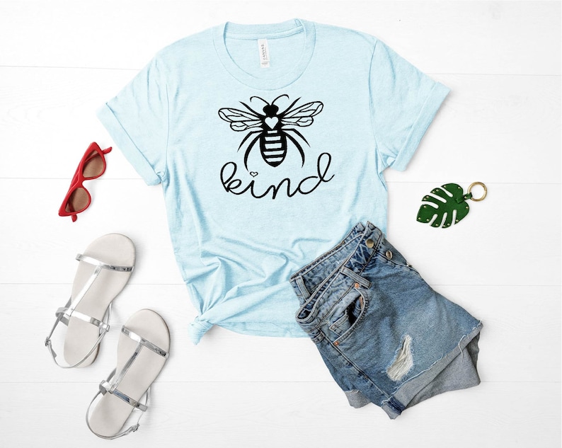 May include: A light blue t-shirt featuring a black bee graphic and the word "kind" printed in black. The bee has a small white heart on its body.