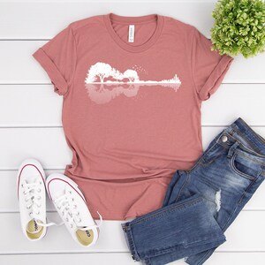 May include: A mauve t-shirt with a white silhouette of a guitar made of trees reflected in water. The t-shirt is laid flat on a white wooden surface with a pair of white sneakers and blue jeans.