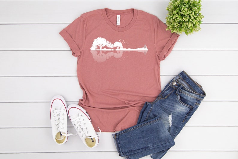 May include: A mauve t-shirt with a white silhouette of a guitar made of trees reflected in water. The t-shirt is laid flat on a white wooden surface with a pair of white sneakers and blue jeans.