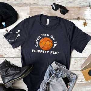 May include: A navy blue t-shirt with a white graphic of a basketball with the text "Catch You On The Flippity Flip".