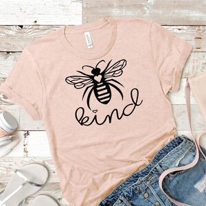 May include: A pink t-shirt featuring a black bee graphic and the word "kind" written in a cursive font. The bee has a small heart on its body.