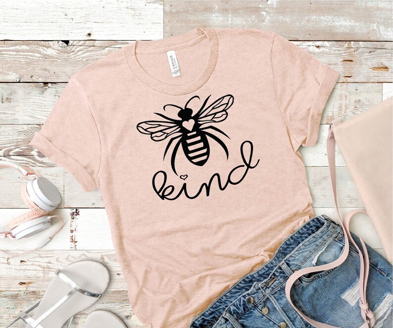 May include: A pink t-shirt featuring a black bee graphic and the word "kind" written in a cursive font. The bee has a small heart on its body.