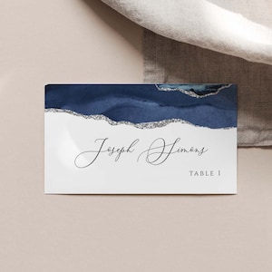 Op de afbeelding: Een wit plaatskaartje met een abstract ontwerp in blauw en zilver. De tekst "Joseph Simons" en "Tafel 1" staat op het kaartje gedrukt.