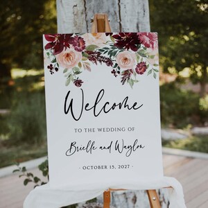 Może przedstawiać: Biały znak powitalny na weselu na drewnianym sztaludze. Znak zawiera kwiatowe akcenty w kolorze bordowym, brzoskwiniowym i zielonym. Słowo "Welcome" jest napisane elegancką czcionką, z imionami Brielle i Waylon oraz datą 15 października 2027.