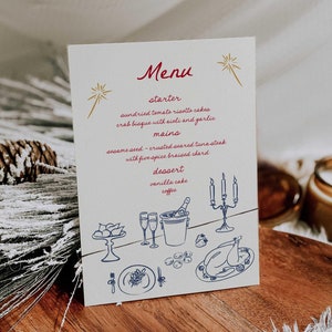 Peut inclure: Une carte de menu blanche avec une illustration dorée et bleue d'un dîner de fête. Le menu comprend des options d'entrée, de plats principaux et de dessert. Le texte sur la carte se lit comme suit : "Menu, entrée, gâteaux de risotto aux tomates séchées, bisque de crabe avec aïoli et ail, plats principaux, steak de thon grillé en croûte de graines de sésame avec du chou braisé aux cinq épices, dessert, gâteau à la vanille, café."